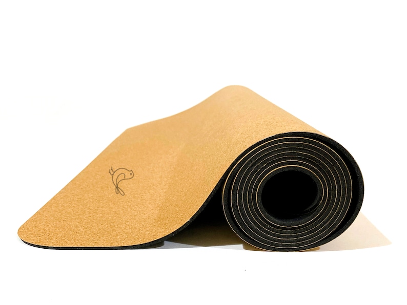May include: A partially rolled yoga mat featuring a light tan, textured cork top surface and a solid black rubberized base. A small, simple black line drawing of a bird is printed on the flat portion of the cork surface near the center.