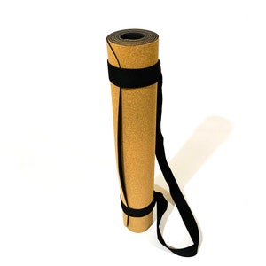 May include: A rolled yoga mat with a tan, cork-textured outer surface and a black inner layer. The mat is secured with two black fabric straps that connect to a long black carrying handle, allowing the mat to be held in a vertical, upright position.