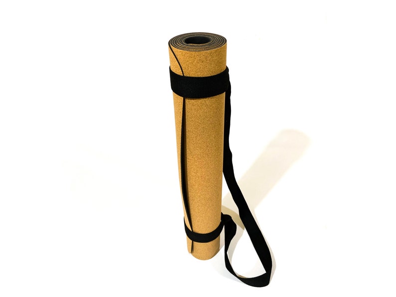 May include: A rolled yoga mat with a tan, cork-textured outer surface and a black inner layer. The mat is secured with two black fabric straps that connect to a long black carrying handle, allowing the mat to be held in a vertical, upright position.