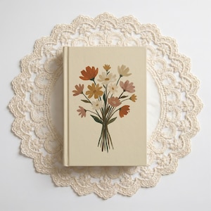 May include: A cream-colored journal with a floral bouquet illustration in shades of orange, pink, yellow, and white. The journal is centered on a cream-colored, lace doily. The flowers are tied together with a green stem.