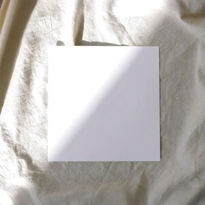 Peut inclure: Une carte carrée d'un blanc uni avec une surface lisse et mate. La carte est posée à plat, présentant une face blanche propre et vierge qui occupe le centre du cadre, avec des bords nets et droits formant une forme géométrique carrée.