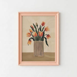 May include: A framed wall art print featuring a bouquet of orange and yellow tulips in a vertically striped cylindrical vase. The artwork is set against a neutral beige background and is enclosed in a light peach-colored wooden frame.