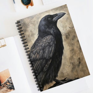 May include: A spiral-bound notebook featuring a detailed illustration of a black raven. The raven is depicted with intricate feather detail against a textured, sepia-toned background. The notebook is open on a white surface.