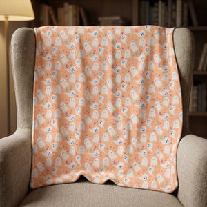 May include: A soft throw blanket featuring a repeating pattern of white ghosts, small colorful stars, and crescent moons set against a solid light orange background. The fabric displays a consistent print of these spectral figures and celestial shapes.