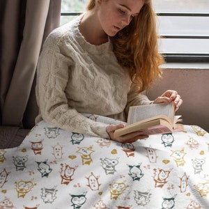 May include: A white throw blanket with a repeating pattern of colorful cartoon characters. The characters are stylized and resemble small animals with large eyes and rounded bodies. The blanket is soft and cozy, perfect for snuggling up on the couch.