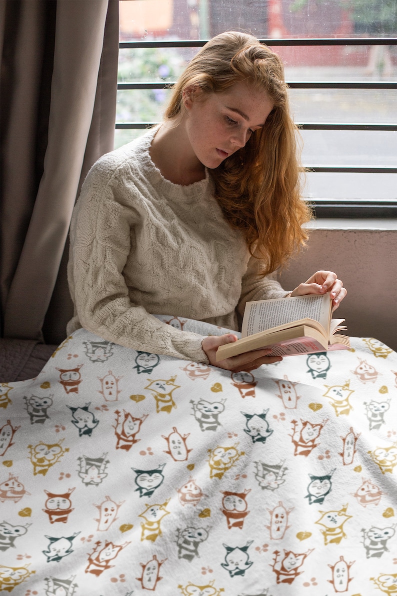 May include: A white throw blanket with a repeating pattern of colorful cartoon characters. The characters are stylized and resemble small animals with large eyes and rounded bodies. The blanket is soft and cozy, perfect for snuggling up on the couch.
