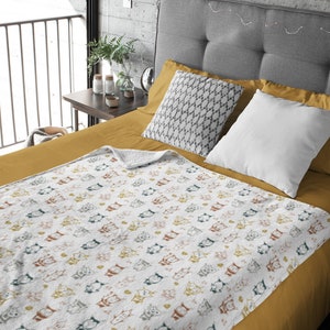 May include: A white throw blanket with a repeating pattern of colorful cartoon cats. The blanket is folded on a mustard yellow bedspread with gray and white pillows.