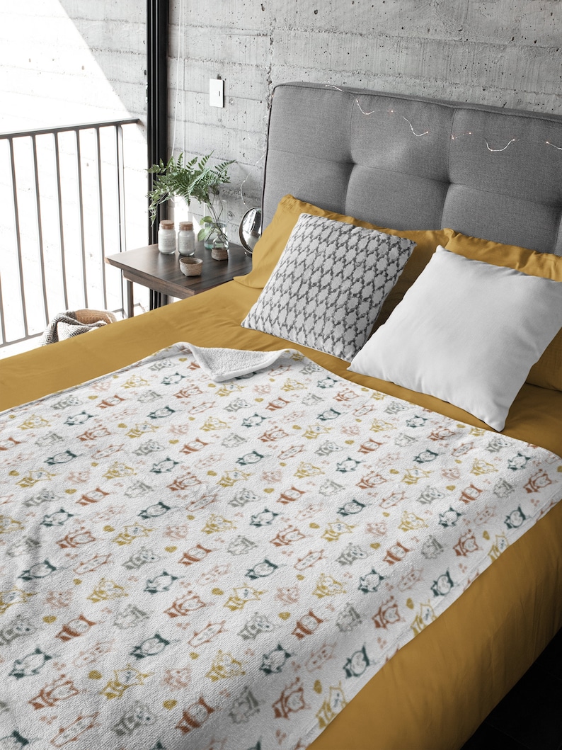 May include: A white throw blanket with a repeating pattern of colorful cartoon cats. The blanket is folded on a mustard yellow bedspread with gray and white pillows.