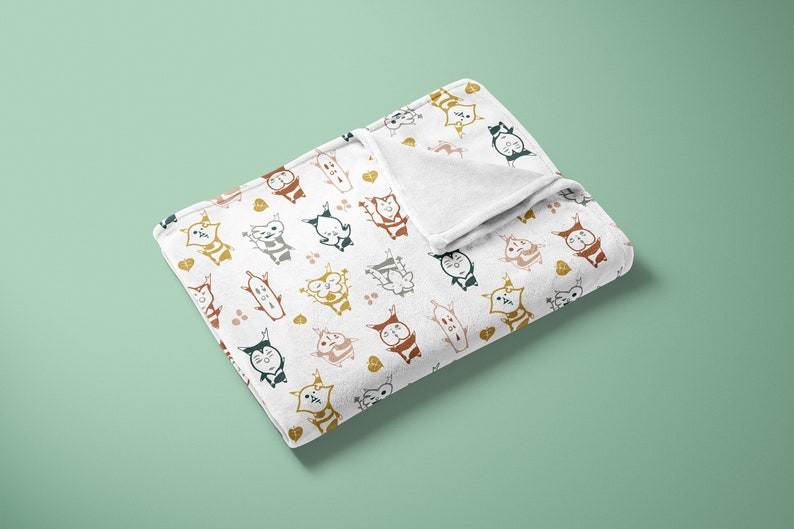 May include: A white fleece blanket with a repeating pattern of colorful cartoon cats. The cats are in various poses and have different facial expressions. The blanket is folded in half and lying on a light green surface.
