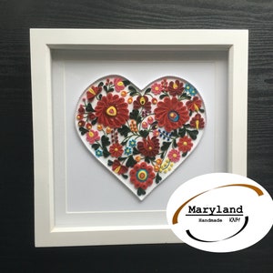 May include: A white frame with a white mat surrounding a heart-shaped paper quilling artwork. The artwork features a red, white, and green floral design. The frame has a brown oval sticker with the text "Maryland Handmade KMH".
