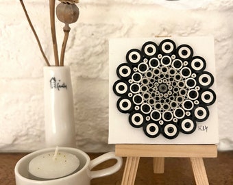 May include: Black and white paper quilling art on a small white canvas with a wooden easel. The art features a circular design with concentric circles. The canvas is labeled "Maryland Handmade KJM".