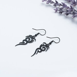 Puede incluir: Un par de pendientes colgantes negros con un diseño en espiral en forma de llama. Los pendientes tienen cierre de gancho y están sobre un fondo blanco con una ramita de flores moradas.