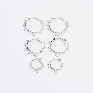 May include: Six silver hoop earrings with a floral design. The earrings are arranged in three pairs of varying sizes, all featuring a detailed, openwork pattern around the hoop's outer edge. The earrings are displayed on a white background.