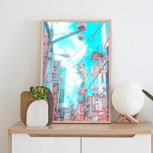 Peut inclure: Une impression encadrée d'une scène de rue colorée au Japon avec un ciel bleu et des bâtiments roses et orange. L'impression est accrochée à un mur blanc au-dessus d'une commode en bois avec une lampe blanche et une plante en pot.