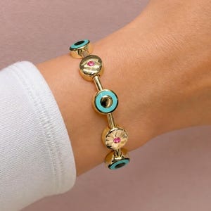 Pode incluir: Uma pulseira de corrente dourada com berloques circulares alternados. Os berloques incluem motivos de olho grego em esmalte turquesa e discos dourados texturizados com uma pequena pedra preciosa redonda de cor rosa no centro.