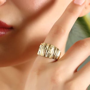 Puede incluir: Un anillo de banda ancha en oro que presenta una serie de ranuras verticales, texturizadas y pulidas en toda la superficie. El anillo tiene un acabado de alto brillo y un perfil ligeramente curvado y abombado que rodea el dedo, creando un aspecto reflectante.