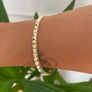 Op de afbeelding: Een goudkleurige armband met een doorlopende rij verhoogde, piramidevormige noppen aan de buitenzijde. Het metaal heeft een gepolijste, reflecterende afwerking en het ontwerp bevat een scharniersluiting die aan de zijkant zichtbaar is.