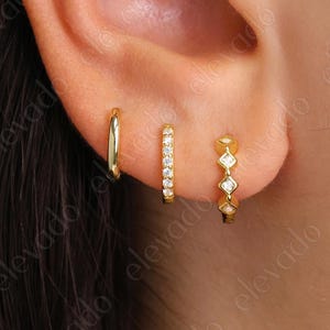 May include: An ear adorned with three gold earrings. The first is a plain hoop, the second a small hoop set with clear stones, and the third a hoop with diamond-shaped detailing.