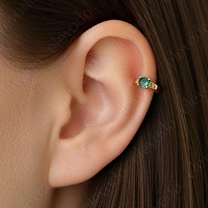 May include: A gold-colored ear cuff with a central oval-shaped green gemstone and smaller green accent stones. The cuff is worn on the ear cartilage, showcasing the jewelry's design and color against the skin.