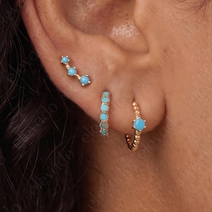 May include: A gold ear climber earring featuring three small round turquoise stones, a gold hoop earring with a row of turquoise stones, and a gold hoop earring with a single turquoise stone, all worn in the earlobe and cartilage piercings.