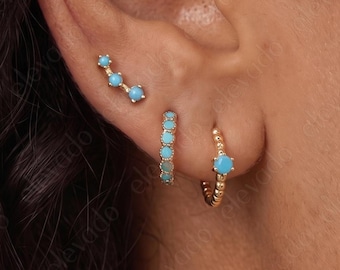 May include: A gold ear climber earring featuring three small round turquoise stones, a gold hoop earring with a row of turquoise stones, and a gold hoop earring with a single turquoise stone, all worn in the earlobe and cartilage piercings.