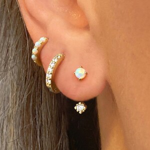 May include: A close-up of a person's ear with three gold earrings. The top earring is a small hoop with white stones. The middle earring is a larger hoop with white stones. The bottom earring is a single white stone with a smaller white stone dangling below.