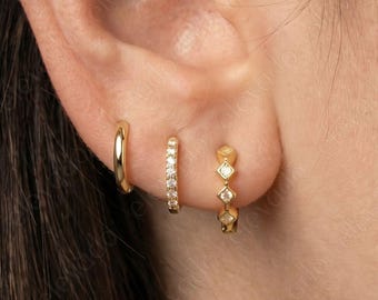 May include: Three gold earrings are displayed on an ear. The first is a small, plain hoop. The second is a small hoop with a line of small, clear stones. The third is a small hoop with three diamond-shaped stones.