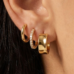 May include: A set of three gold-toned hoop earrings worn in a single ear. The first is a plain thin hoop, the second is a thin hoop encrusted with small clear stones, and the third is a wider, smooth, polished gold-toned hoop earring.