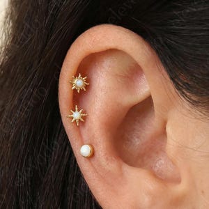 Może przedstawiać: Zbliżenie ucha ozdobionego trzema złotymi kolczykami. Dwa mają kształt gwiazdy z białymi akcentami, a trzeci to mały, okrągły biały kolczyk. Kolczyki są umieszczone na tle ciemnych włosów i skóry.