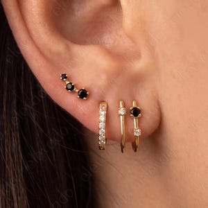 May include: A set of four gold earrings worn in a single ear. The set includes a curved stud with three black stones, a small hoop with clear stones, a thin plain gold hoop, and a small hoop featuring one black stone and one clear stone.