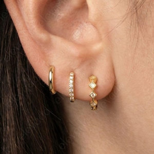 Peut inclure: Trois boucles d'oreilles dorées sont présentées sur une oreille. La première est un petit anneau simple. La seconde est un petit anneau avec une ligne de petites pierres claires. La troisième est un petit anneau avec trois pierres en forme de diamant.