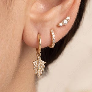 Peut inclure: Un ensemble de trois boucles d'oreilles dorées portées au lobe et au cartilage. La boucle inférieure est un anneau avec une breloque en forme de feuille, celle du milieu est un petit anneau pavé, et celle du haut est un clou incurvé, toutes serties.