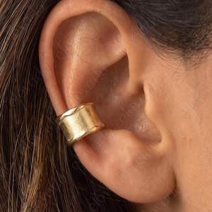 May include: A gold-toned ear cuff featuring a smooth, wide band design with slightly raised edges. The piece is worn on the outer cartilage of the ear, showcasing a polished metallic finish that reflects light across its curved surface.