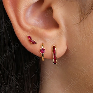 Könnte beinhalten: Nahaufnahme eines Ohrs mit drei goldenen Ohrringen. Die Ohrringe sind alle mit roten Edelsteinen besetzt. Die Ohrringe sind ein kleiner Stecker, ein größerer Stecker und ein Creolen.