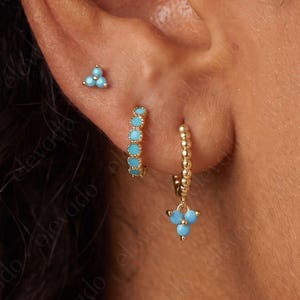 May include: A gold stud earring featuring three small turquoise stones in a triangular cluster, a gold hoop earring with turquoise stones, and a gold hoop earring with a dangling three-stone turquoise charm worn in an earlobe.