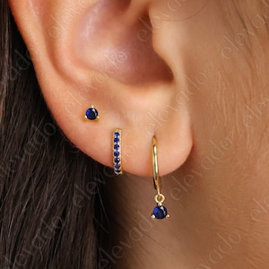 May include: A close-up of a person's ear with three gold earrings. The earrings are all small and feature blue gemstones. The top earring is a simple stud, the middle earring is a small hoop with a row of blue gemstones, and the bottom earring is a hoop with a single blue gemstone dangling from it.