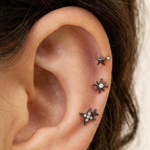 May include: An ear with three star-shaped earrings. The top earring is a small, solid star. The middle earring is a starburst design. The bottom earring is a star with multiple small, clear stones. All earrings are dark in colour.