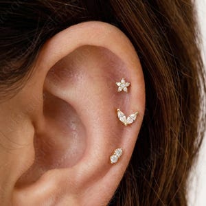 May include: A close-up of an ear adorned with three gold earrings. The top earring is a small flower, the middle is a curved design, and the bottom is a double-stone stud. All earrings feature clear, sparkling stones.
