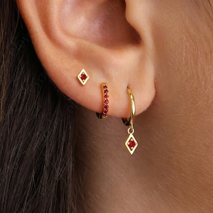 May include: A close-up of a person's ear with three gold earrings. The earrings are all set with small red gemstones. The top earring is a small square stud, the middle earring is a small hoop with red gemstones around the edge, and the bottom earring is a hoop with a dangling square charm set with red gemstones.