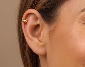 May include: A small gold-toned earring shaped like a hummingbird, positioned in the upper cartilage of the ear. The earring features a polished metallic finish and a detailed silhouette of a bird with wings slightly extended in a flying pose.