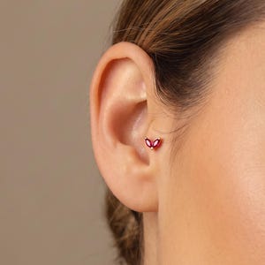 Peut inclure: Une petite boucle d'oreille en or ornée de deux pierres précieuses rouges taille marquise disposées en forme de V. Le bijou est placé dans la conque de l'oreille, présentant un design délicat avec une monture en métal poli.