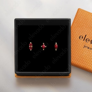 May include: A black jewelry box with three red gemstone earrings. The earrings are displayed on a black velvet interior. An open orange box with the text "elevado jewelry" is next to the jewelry box.