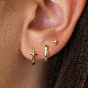 Puede incluir: Una oreja con tres pendientes de oro. Los pendientes son una pequeña estrella, un aro pequeño y un aro más grande. Los aros son lisos y tienen un diseño de estrella.