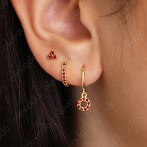May include: Three gold earrings with red gemstones. The earrings are a small triangle stud, a small hoop with red gemstones, and a larger hoop with a dangling charm that is also set with red gemstones.