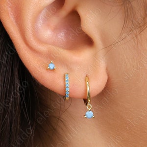 May include: A close-up of a person's ear with three gold earrings. The earrings are all small and have blue stones. The top earring is a stud, the middle earring is a hoop, and the bottom earring is a dangle.