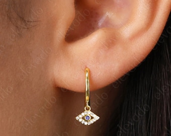 May include: Gold hoop earrings with a dangling evil eye charm. The charm is set with small white stones and a blue center stone.