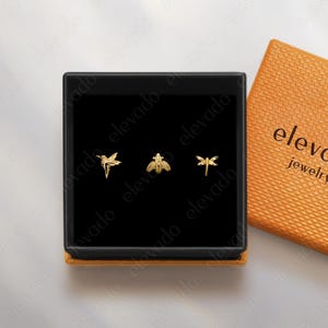 May include: A black jewelry box with three gold-colored stud earrings shaped like a bird, bee, and dragonfly. The box is open, revealing the earrings against a black interior. An orange textured box with the text "elevado jewelry" is next to it.