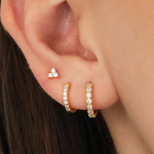 Pode incluir: Close-up de uma orelha adornada com três peças de joalharia. Um pequeno brinco de ouro com três pedras transparentes está posicionado no topo. Abaixo, um pequeno brinco de argola de ouro com pedras transparentes é visível. Outro brinco de argola de ouro com pedras transparentes está abaixo.