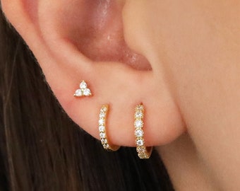 May include: Close-up of a person's ear adorned with three pieces of jewelry. A small gold stud earring with three clear stones is positioned at the top. Below, a small gold hoop earring with clear stones is visible. Another gold hoop earring with clear stones is below.