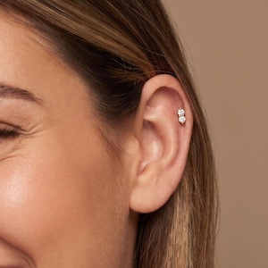 May include: A gold-toned cartilage earring featuring two small, clear, round-cut stones set vertically in a metal post. The jewelry is positioned in the upper ear cartilage, showcasing a simple, reflective design with a polished finish.